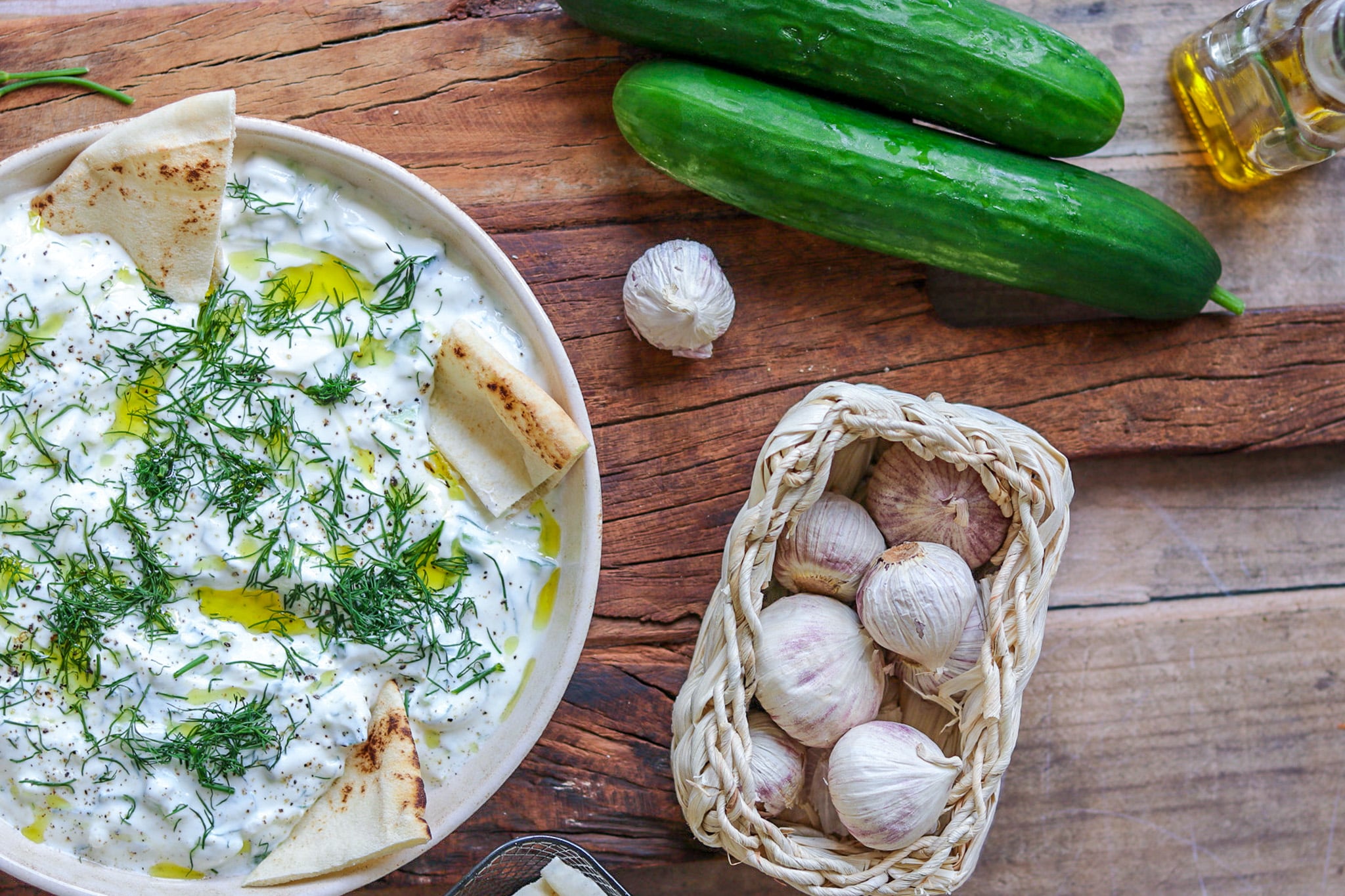 Tstatsiki mit griechischem Joghurt serviert in einer hellen Schale, mit Deko auf einme Holzbrett, Knoblauch und Gurken