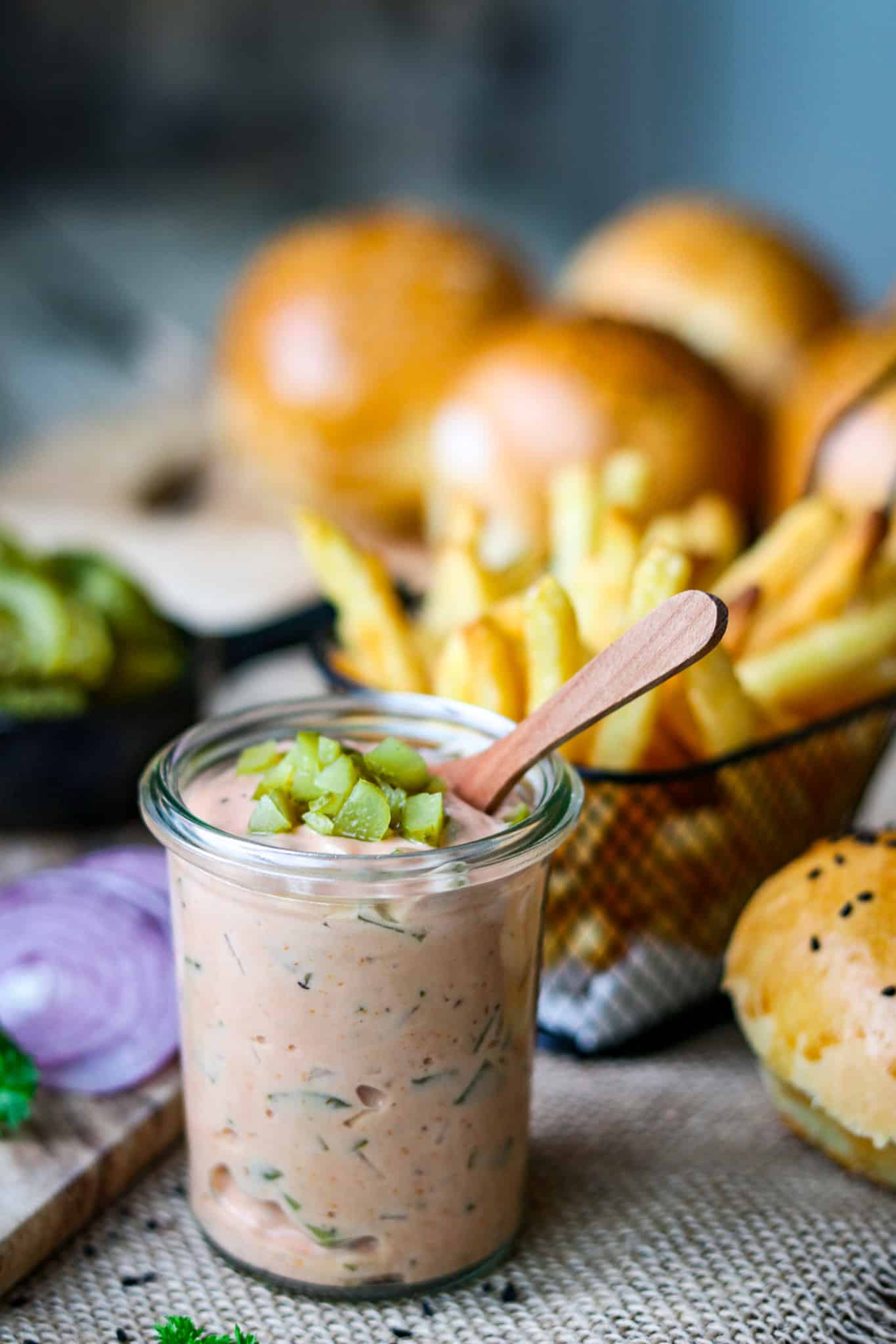 Burger Soße in einem kleinen Glas und einem kleinen Holzlöffel in der Soße, im Hintergrund erkennt man Burger und Pommes und rote Zwiebelringe, alles steht auf einem rustikalen Holzuntergrund.