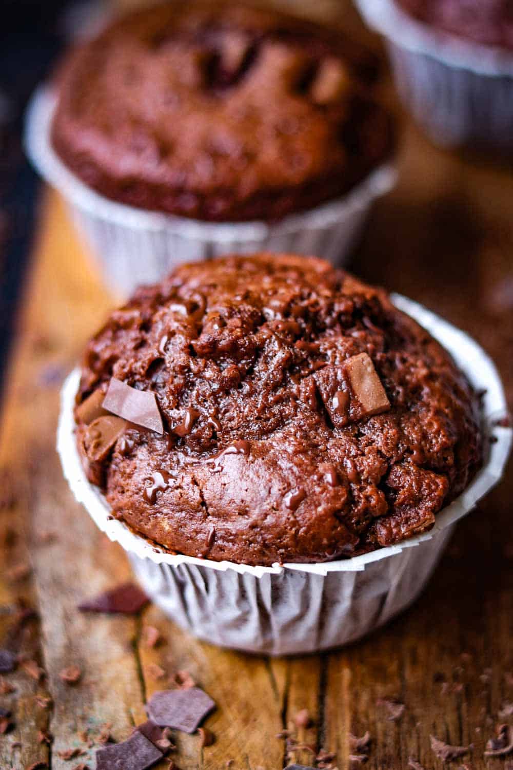 Double Chocolate Muffins mit Papier und Schokoladenstückchen auf einem rustikalen Brett und dunklem Untergrund